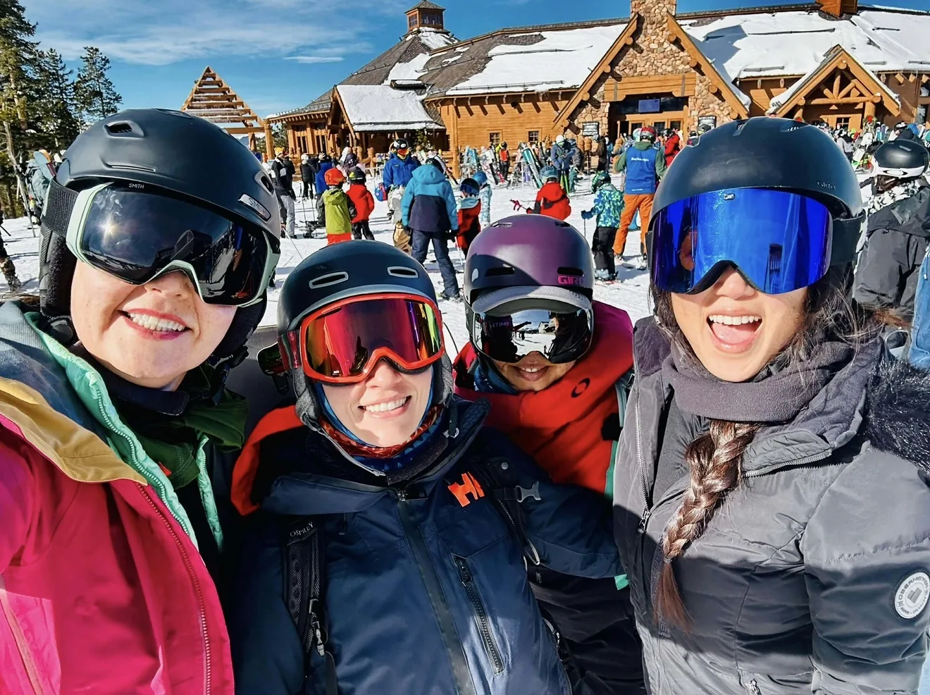 Four smiling friends in ski helmets and goggles at a snowy mountain lodge